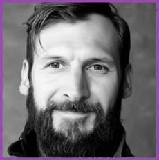 Close-up black-and-white portrait of a man with a full beard and short hair, looking at the camera, framed by a purple border.