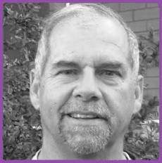 Close-up grayscale portrait of a smiling man with short hair and a goatee, framed by a purple border.